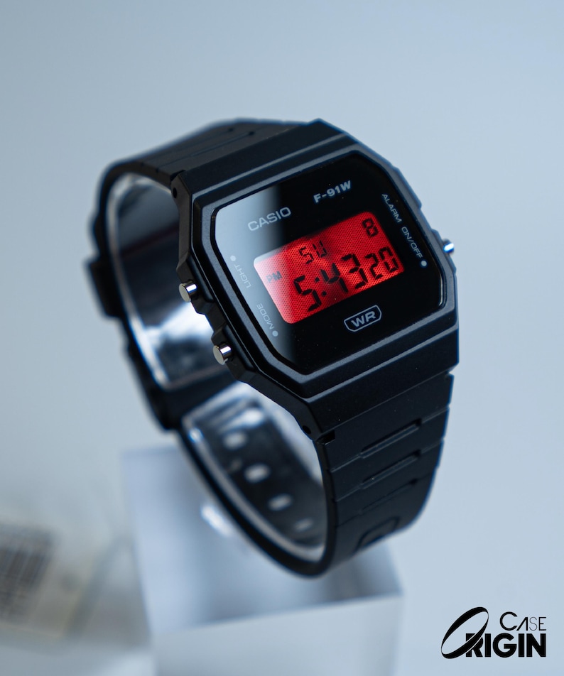 May include: A black Casio F-91W digital watch with a rectangular face and a black resin strap. The red digital display shows the time. The watch has the text "CASIO" and "F-91W" printed on the face.