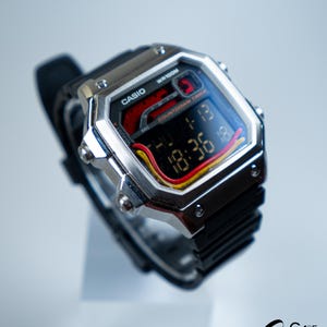Puede incluir: Un reloj digital Casio plateado con una correa negra. La esfera del reloj muestra la hora y la fecha en formato digital, con detalles rojos, amarillos y negros. El reloj está etiquetado como "WR100M" y "Casio".