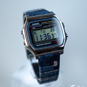 May include: A Casio digital watch with a blue and silver case and a blue denim strap. The watch face displays the time, day, and date. The watch is water-resistant and has an alarm and chronograph functions.