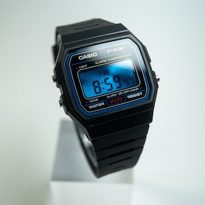 May include: Black Casio F-91W digital watch with a blue display showing the time 8:59:44. The watch has a black resin strap and is water-resistant. The watch face includes the Casio logo and the text "ALARM CHRONOGRAPH".