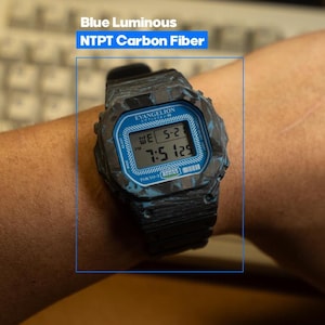 May include: A black and blue digital watch with a blue face and the text "EVANGELION" and "TOKYO-3" printed on the watch face. The watch has a carbon fiber design and the text "Blue Luminous NTPT Carbon Fiber" is above the watch.