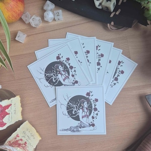 Könnte beinhalten: Eine Sammlung quadratischer Kunstdrucke mit einer Frau mit einem dunklen Kreis und Rosendesign. Die Drucke sind auf einer Holzoberfläche angeordnet, mit Würfeln, Pflanzen und einer Flaschen-Potion im Hintergrund.