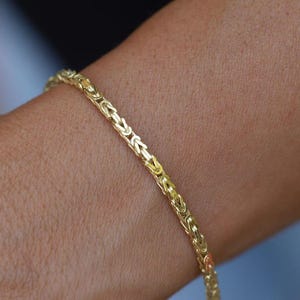 Puede incluir: Una pulsera de cadena bizantina dorada que presenta una serie de eslabones metálicos entrelazados y tejidos que crean una apariencia cilíndrica texturizada. La pulsera tiene un acabado pulido y reflectante, luciendo un patrón intrincado en la muñeca.