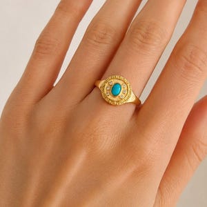 Könnte beinhalten: Ein goldfarbener Ring mit einem zentralen ovalen Türkisstein, der von einem texturierten Rand mit kleinen, eingelassenen klaren Steinen umgeben ist. Die kreisförmige Vorderseite des Rings zeigt ein detailliertes Muster am Rand auf einem polierten Goldband.