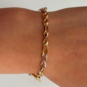Puede incluir: Una pulsera de cadena de oro con una serie de eslabones entrelazados en forma de infinito. El diseño alterna entre eslabones de oro amarillo pulido y un eslabón plateado texturizado, creando un patrón de bucle continuo alrededor de la muñeca.