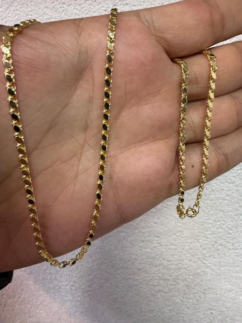 May include: A gold-colored chain necklace with a delicate, repeating oval link design. The necklace is displayed on a hand, showcasing its length and the intricate pattern of the links. The clasp is visible at the end of the chain.