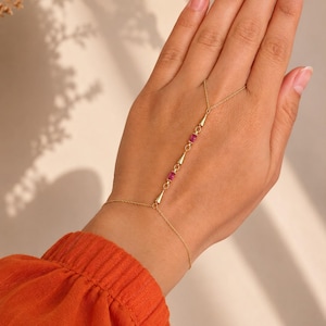 Könnte beinhalten: Ein Handkettchen aus Gold mit einer feinen Kette, die ein Armband mit einer Fingerschlaufe verbindet. Das Design umfasst zwei kleine rechteckige rosafarbene Edelsteine und drei kegelförmige Goldakzente entlang des zentralen Kettenabschnitts.