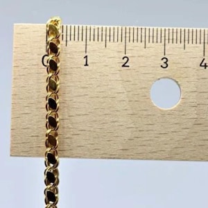 May include: A gold-colored chain with an oval link design is positioned next to a wooden ruler. The ruler shows measurements in inches, with the chain extending from the zero-inch mark. The chain's links are closely connected, creating a continuous pattern.