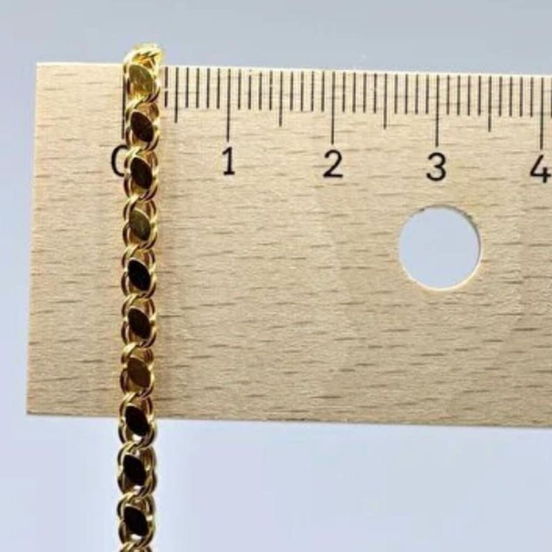 May include: A gold-colored chain with an oval link design is positioned next to a wooden ruler. The ruler shows measurements in inches, with the chain extending from the zero-inch mark. The chain's links are closely connected, creating a continuous pattern.