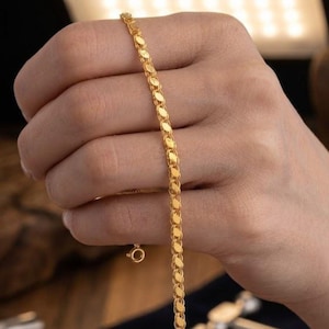 May include: A gold-colored bracelet with an oval link design. The bracelet is held in a hand, showcasing its delicate chain and clasp. The bracelet's links are closely connected, creating a continuous pattern. The gold color gives the bracelet a classic and elegant look.