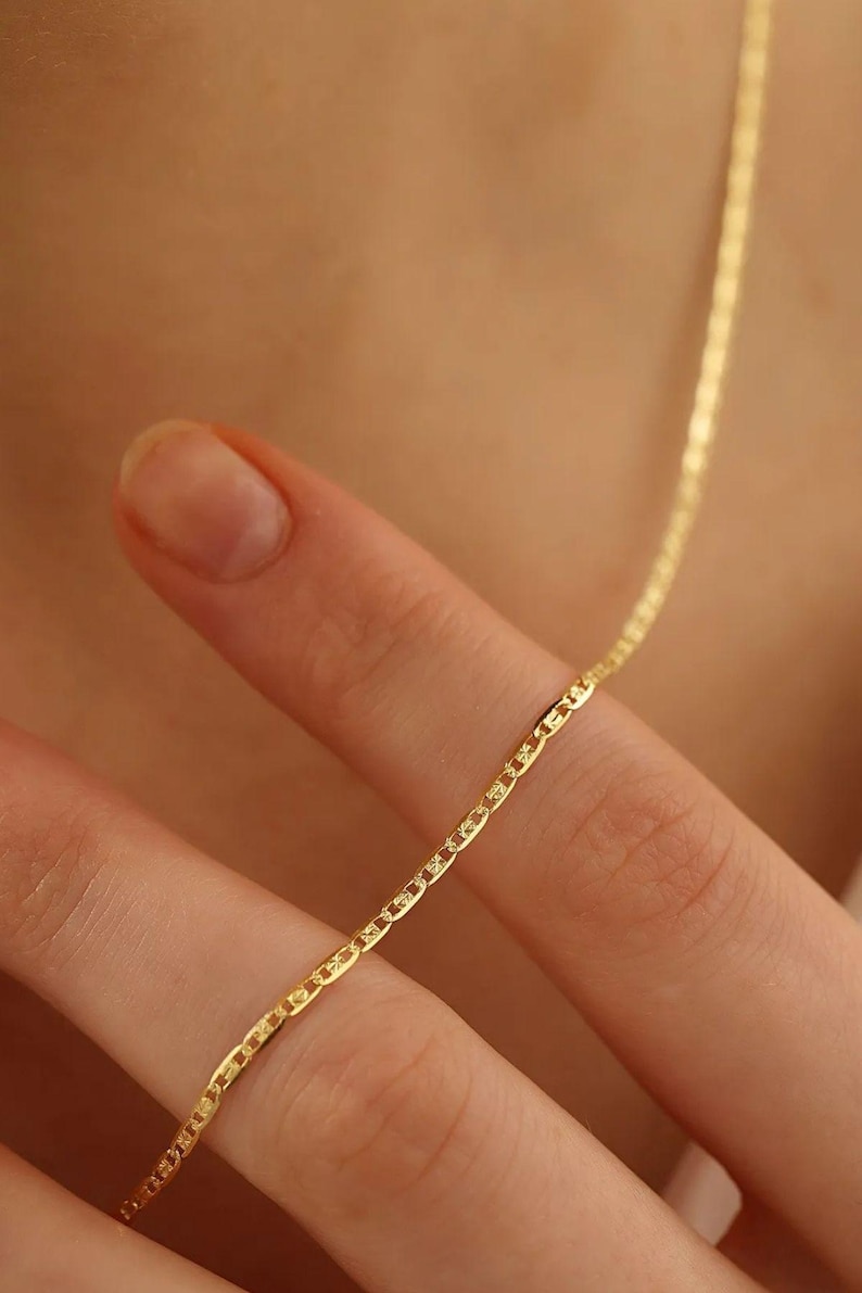 May include: A gold chain necklace with a delicate, mariner link design. The chain is shown wrapped around a finger.