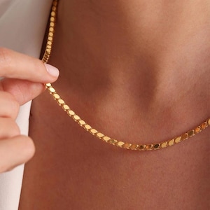 May include: A gold-colored necklace with a delicate, oval-link chain. The necklace is shown against a skin tone background, with a hand gently holding a portion of the chain. The chain is a bright, shiny gold.