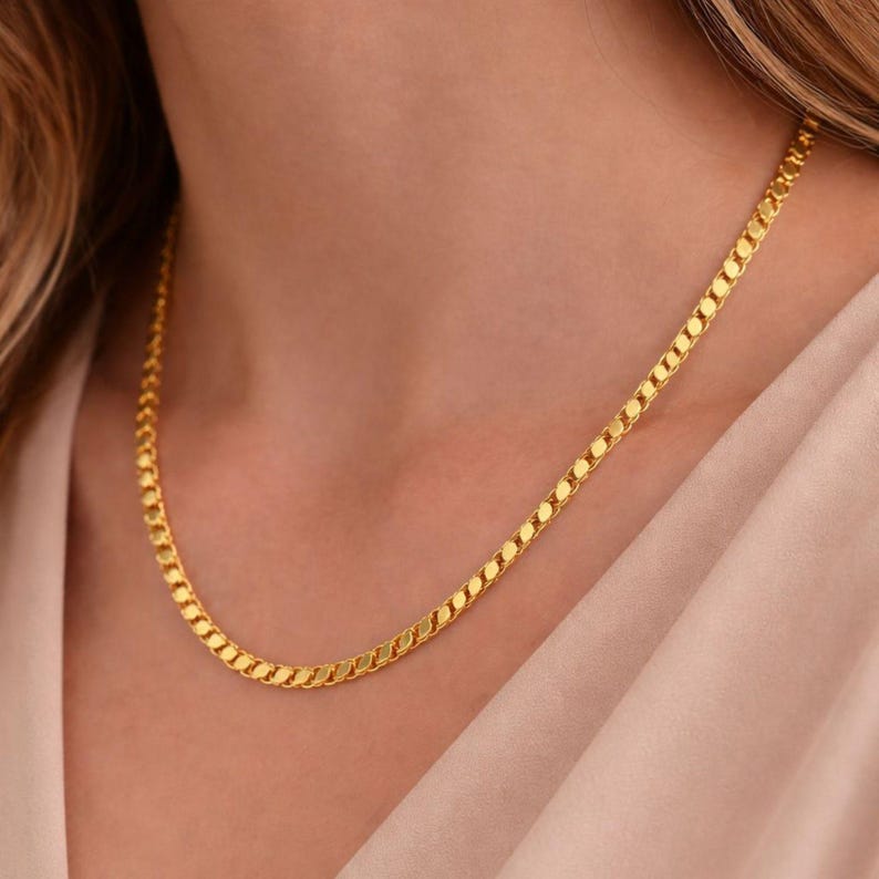 May include: A gold-colored chain necklace with a flat curb link design. The necklace is worn around the neck and rests on a light-colored fabric. The chain links are closely connected, creating a smooth, continuous appearance.