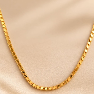 May include: A gold-colored necklace with a delicate, flat chain design. The chain is composed of small, oval-shaped links that catch the light, creating a shimmering effect. The necklace is displayed against a soft, neutral background.