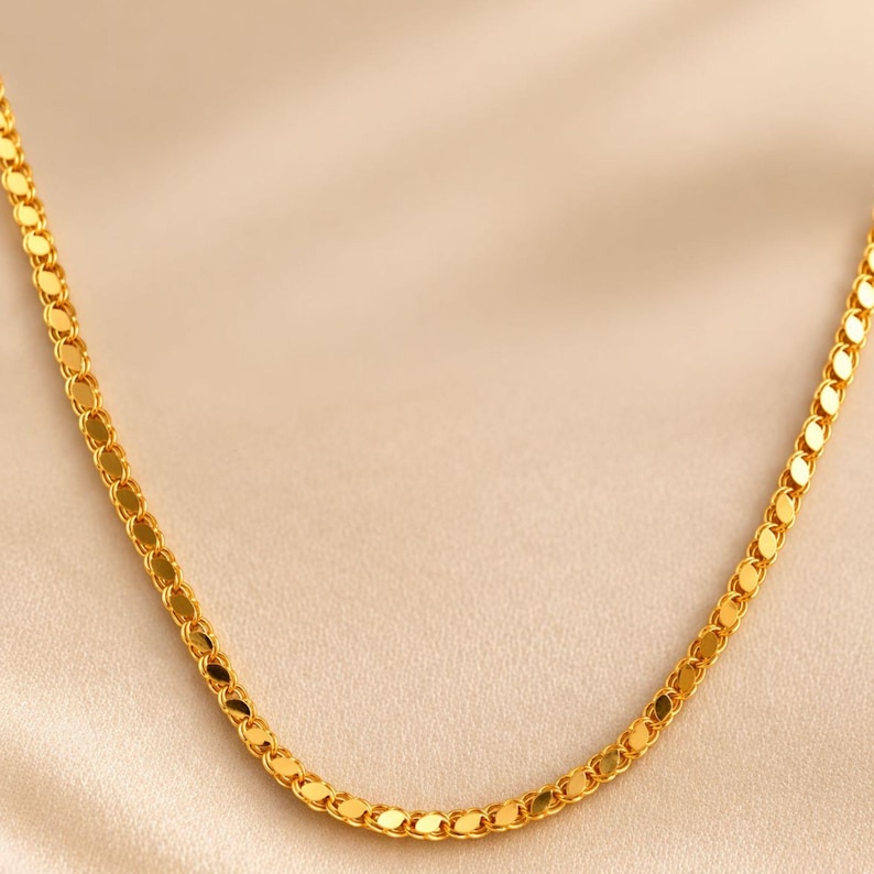 May include: A gold-colored necklace with a delicate, flat chain design. The chain is composed of small, oval-shaped links that catch the light, creating a shimmering effect. The necklace is displayed against a soft, neutral background.