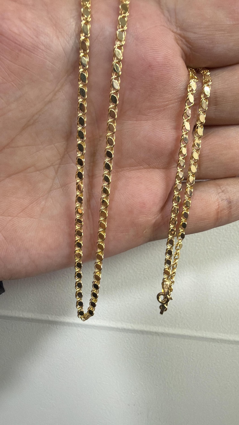 May include: A delicate gold necklace with a repeating oval link design. The chain is displayed against a neutral background, showcasing its bright, reflective surface. The necklace is a classic jewelry piece.