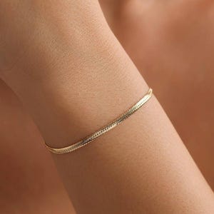 May include: A gold-colored herringbone bracelet is worn on a person's arm. The bracelet is made of small, interlocking links that create a flat, flexible band. The bracelet is a simple, elegant piece of jewelry.