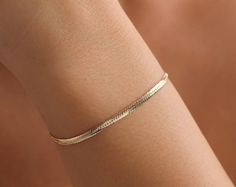 Könnte beinhalten: Ein goldfarbenes Fischgrätenarmband wird an einem Arm getragen. Das Armband besteht aus kleinen, ineinandergreifenden Gliedern, die ein flaches, flexibles Band bilden. Ein schlichtes, elegantes Schmuckstück.