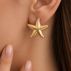 Pode incluir: Um brinco de pino dourado em forma de estrela-do-mar de cinco pontas. A superfície da estrela-do-mar apresenta uma borda texturizada com contas ao longo de cada um dos seus cinco braços, com um centro liso e polido que reflete a luz.