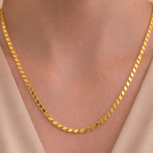 May include: A gold-colored necklace with a flat, oval-link chain. The necklace is draped around a person's neck, with the chain lying flat against the skin. The background is a soft, neutral color.