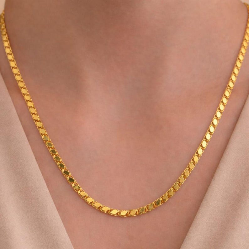 May include: A gold-colored necklace with a flat, oval-link chain. The necklace is draped around a person's neck, with the chain lying flat against the skin. The background is a soft, neutral color.