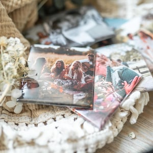 Könnte beinhalten: Eine Sammlung glänzender Fotoabzüge mit verschiedenen Szenen im Freien, darunter eine Gruppe von Menschen am Lagerfeuer. Die Fotos liegen auf einer weißen Spitzendecke mit Trockenblumen und einem beigen Stoff.