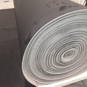 Puede incluir: Un rollo de material de espuma grueso de color gris oscuro con un borde gris claro. El rollo está sobre una superficie blanca. El material parece nuevo y sin usar. La espuma probablemente se utiliza para aislamiento o acolchado.
