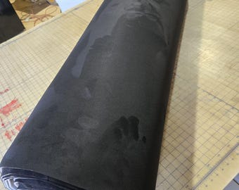 May include: A large roll of matte black fabric with a smooth, uniform surface texture. The material is tightly wound into a cylindrical shape, revealing a light gray underside at the end of the roll. The fabric rests on a flat surface.