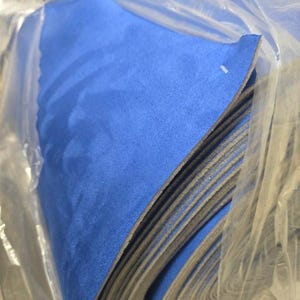 Puede incluir: Una pila de hojas de tela azules, posiblemente fieltro o ante, es visible dentro de una bolsa de plástico transparente. La tela tiene una textura suave y un color azul vibrante. Los bordes de las hojas son grises.