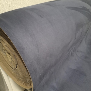Puede incluir: Un rollo de tela azul con una textura suave similar a la gamuza. La tela tiene una apariencia veteada con remolinos y patrones azul oscuro. El rollo está parcialmente desenrollado, revelando el material de respaldo beige.