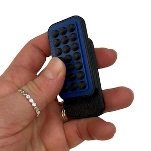 May include: A blue and black rectangular device with a grid of black circular bumps. The device is held in a hand, and a silver ring is visible on the finger. The device appears to be made of plastic.