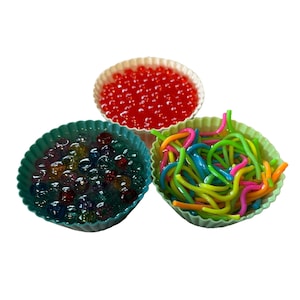 May include: Three colorful sensory toys in silicone cupcake molds. One mold contains red water beads, another has clear and colorful marbles, and the third holds a mix of colorful, bendable, and squiggly noodles. The molds are white, teal, and green.