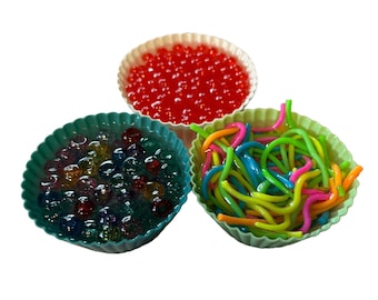 May include: Three colorful sensory toys in silicone cupcake molds. One mold contains red water beads, another has clear and colorful marbles, and the third holds a mix of colorful, bendable, and squiggly noodles. The molds are white, teal, and green.