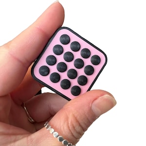 May include: A pink square fidget toy with a black border and 16 black circular buttons. The toy is held in a hand against a white background. The buttons are arranged in a grid pattern.