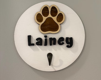 May include: White round wooden sign with a brown paw print and the name "Lainey" in black lettering. A small metal hook is attached to the bottom of the sign.