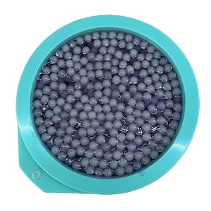May include: A circular, turquoise container filled with small, grey spheres. The spheres are densely packed and appear to be submerged in a clear liquid. The container has a small, protruding tab on the side.