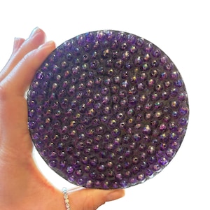 Puede incluir: Un posavasos redondo y morado cubierto de pequeñas cuentas iridiscentes. El posavasos se sostiene en una mano sobre un fondo blanco. Las cuentas reflejan la luz, creando un efecto brillante.