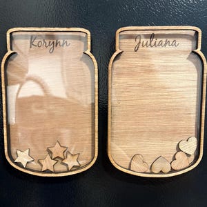 May include: Two wooden mason jar-shaped signs with clear glass inserts. The sign on the left has the name "Korynn" engraved on it and has wooden stars inside the glass. The sign on the right has the name "Juliana" engraved on it and has wooden hearts inside the glass.