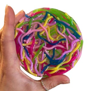 May include: A round, colorful resin coaster filled with vibrant, squiggly, worm-like shapes. The coaster features a mix of bright colors, including pink, yellow, green, blue, and purple. The coaster is held in a hand against a white background.