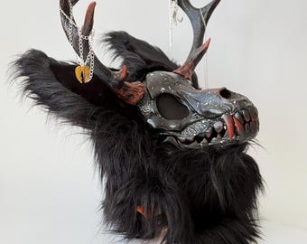 May include: A fantasy mask in black and grey, featuring faux fur and antlers. The mask has a skull-like design with red detailing on the teeth and antlers. A silver chain with a pendant hangs from the antlers. Ideal for cosplay or costume events.