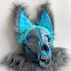 May include: A gray and blue skull-shaped mask with a furry gray and blue trim. The mask has blue accents and mesh eye coverings. The ears are blue with gray fur. The mask is designed to be worn.