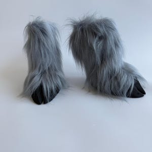 May include: A pair of gray furry boots with black hoof-shaped soles. The boots have long, shaggy fur and are designed to resemble animal feet. The boots are set against a white background.