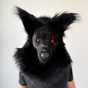 Puede incluir: Una máscara de lobo negro con ojos rojos y una cicatriz. La máscara está hecha de piel sintética y tiene orejas grandes y puntiagudas. La persona que lleva la máscara lleva una camiseta negra.