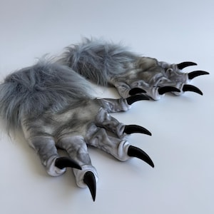 Puede incluir: Un par de guantes de piel sintética gris y blanca con largas garras negras. Los guantes tienen una textura peluda y están diseñados para parecerse a las patas de un lobo o monstruo. Las garras son curvas y puntiagudas.