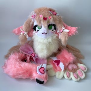 May include: A pink and white furry plush toy with a heart-shaped nose and green eyes. The toy has pink bows in its hair and is wearing a pink and white outfit with wings and hearts.