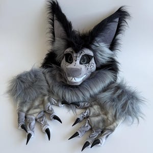 Puede incluir: Una máscara de criatura peluda gris y negra con mangas y guantes con garras a juego. La máscara tiene orejas grandes, una cara detallada con un hocico blanco y garras negras. El diseño incluye una telaraña y otros detalles.