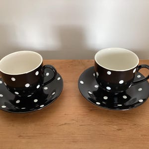 May include: Two black ceramic tea cups with matching saucers, each featuring a white polka dot pattern. The cups have a smooth, glossy finish with a white interior, a curved handle on the side, and a circular base that rests on the matching saucer.