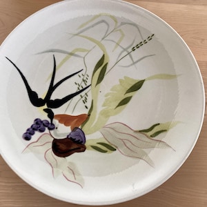 May include: A white ceramic plate with a hand-painted design of a bird, leaves, and grapes. The design is in shades of green, brown, black, and purple.