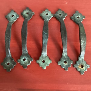 May include: Five antique bronze drawer pulls with a decorative, ornate design. Each pull features a curved handle and a diamond-shaped base with a central hole for mounting. The pulls have a weathered, aged appearance.