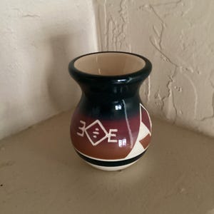 May include: A small, handcrafted ceramic vase with a dark teal top and a beige interior. The body features a geometric design in white, brown, and red. A black stripe separates the body from the white base.